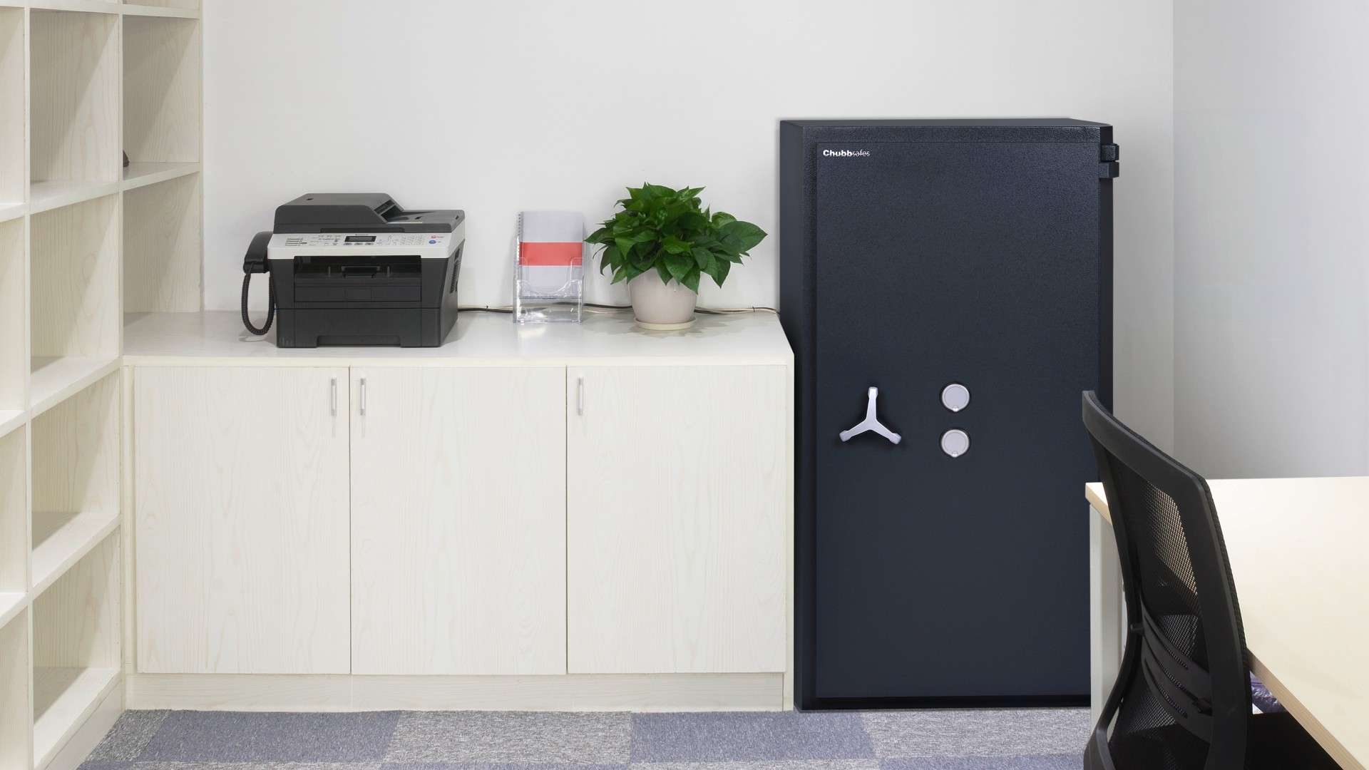 Cassaforte nera in un ambiente domestico o ufficio, posizionata accanto a una stampante e una pianta